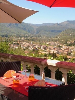 A table with a red tablecloth, a bottle, and glasses, overlooking a town with mountains in the background. - Auberge Provencale (Sospel)