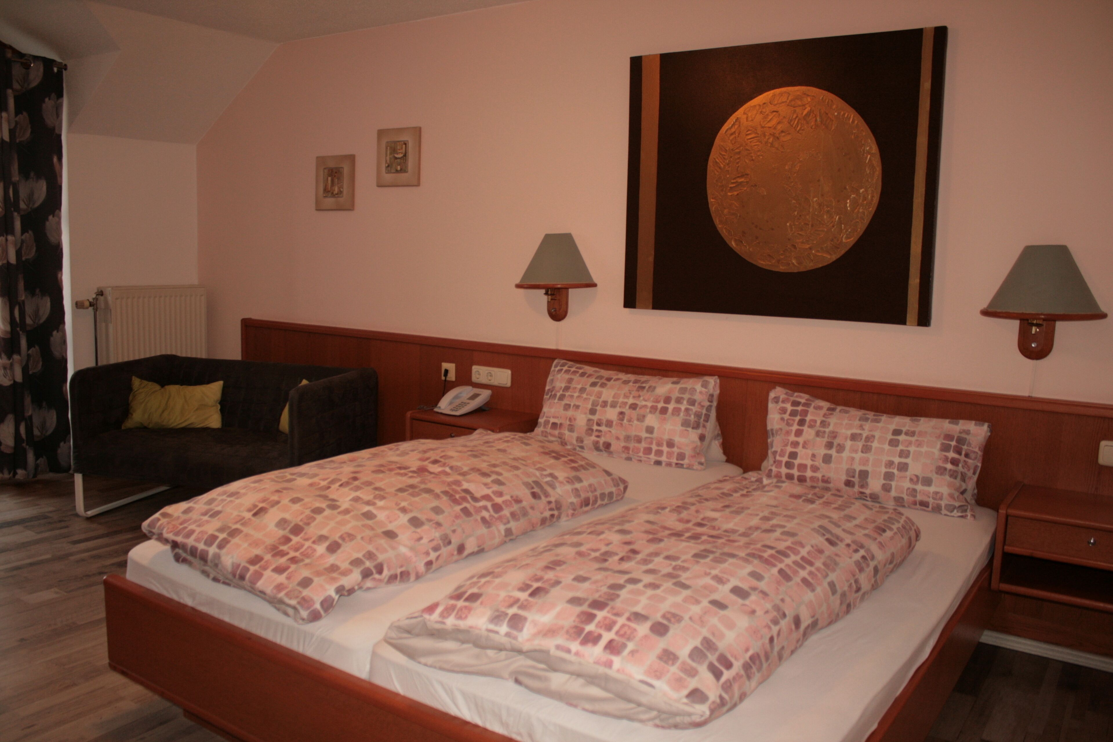 A double bed with a patterned bedspread, a bedside table with a lamp, a nightstand with a phone, a dark-colored sofa, and a large circular artwork on the wall.