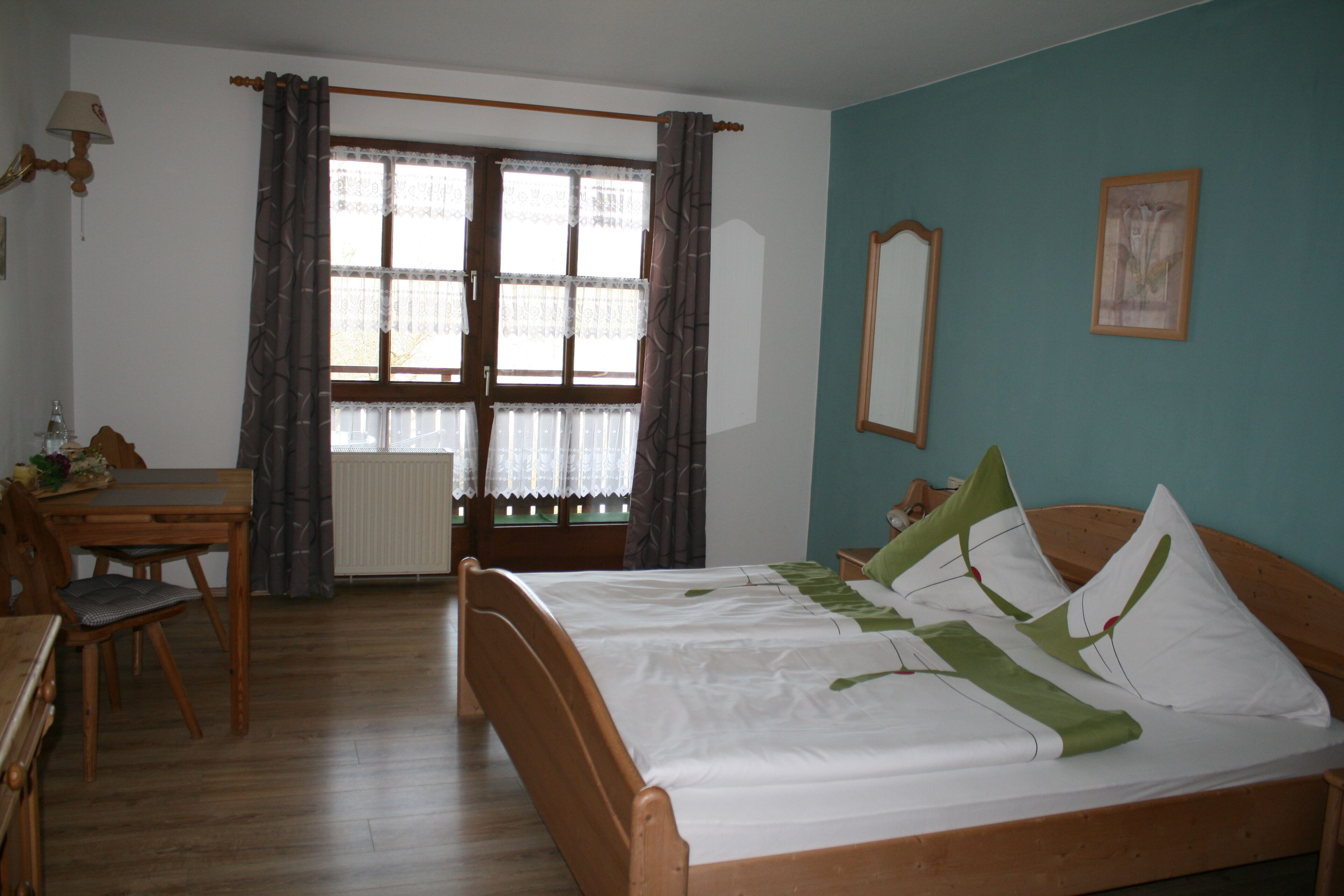 Ein Zimmer mit einem hölzernen Bett, einem Schreibtisch aus Holz, einem Stuhl, einem Fenster mit Vorhängen und einem an der Wand befestigten Spiegel.