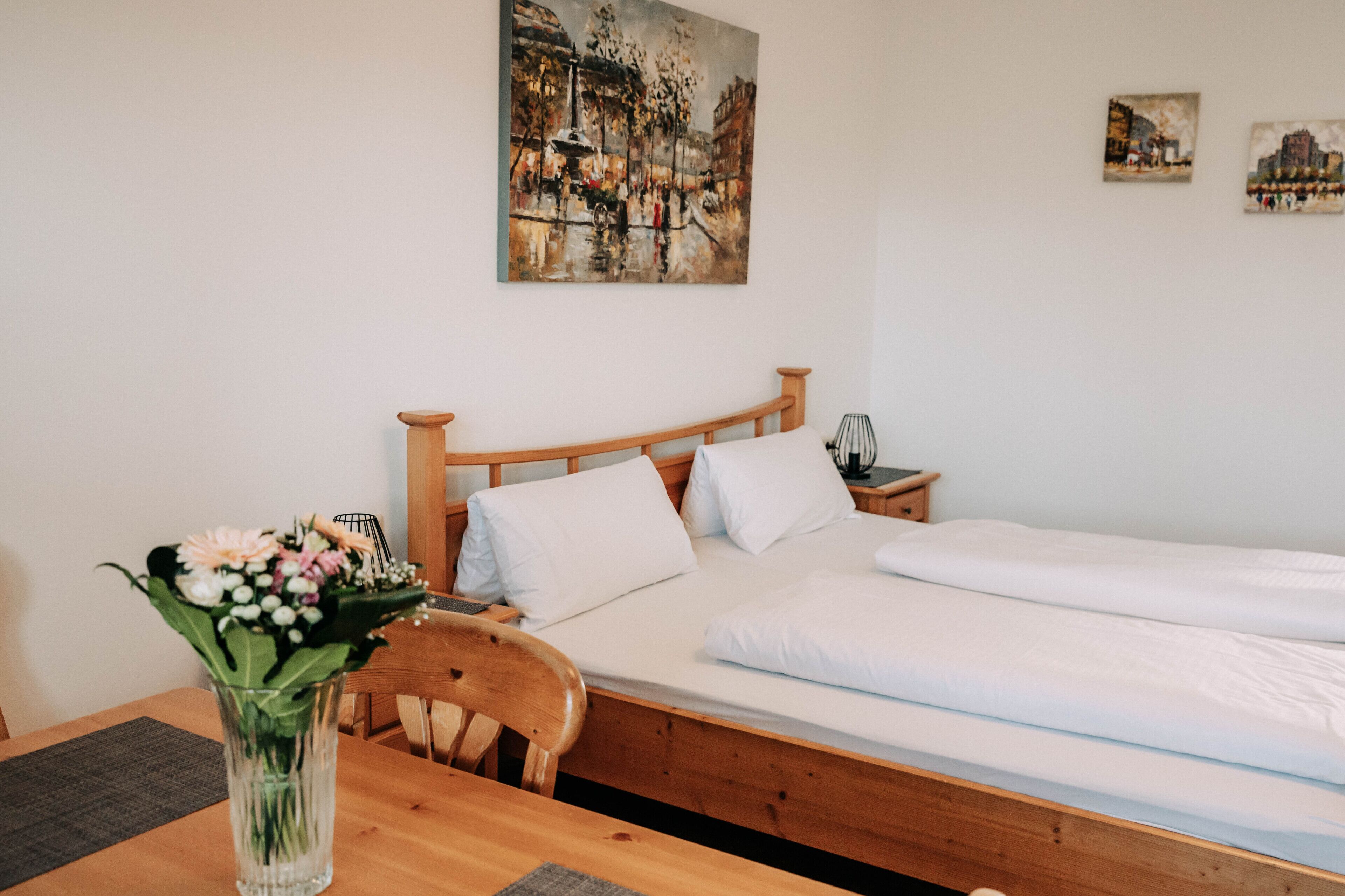 A room with a wooden bed, white bedding, a wooden nightstand, a vase with flowers, and a painting on the wall.
