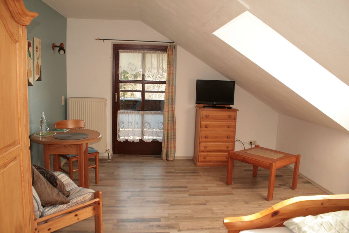 A cozy room with a wooden bed, a small wooden table, a television on a dresser, and a window with curtains.