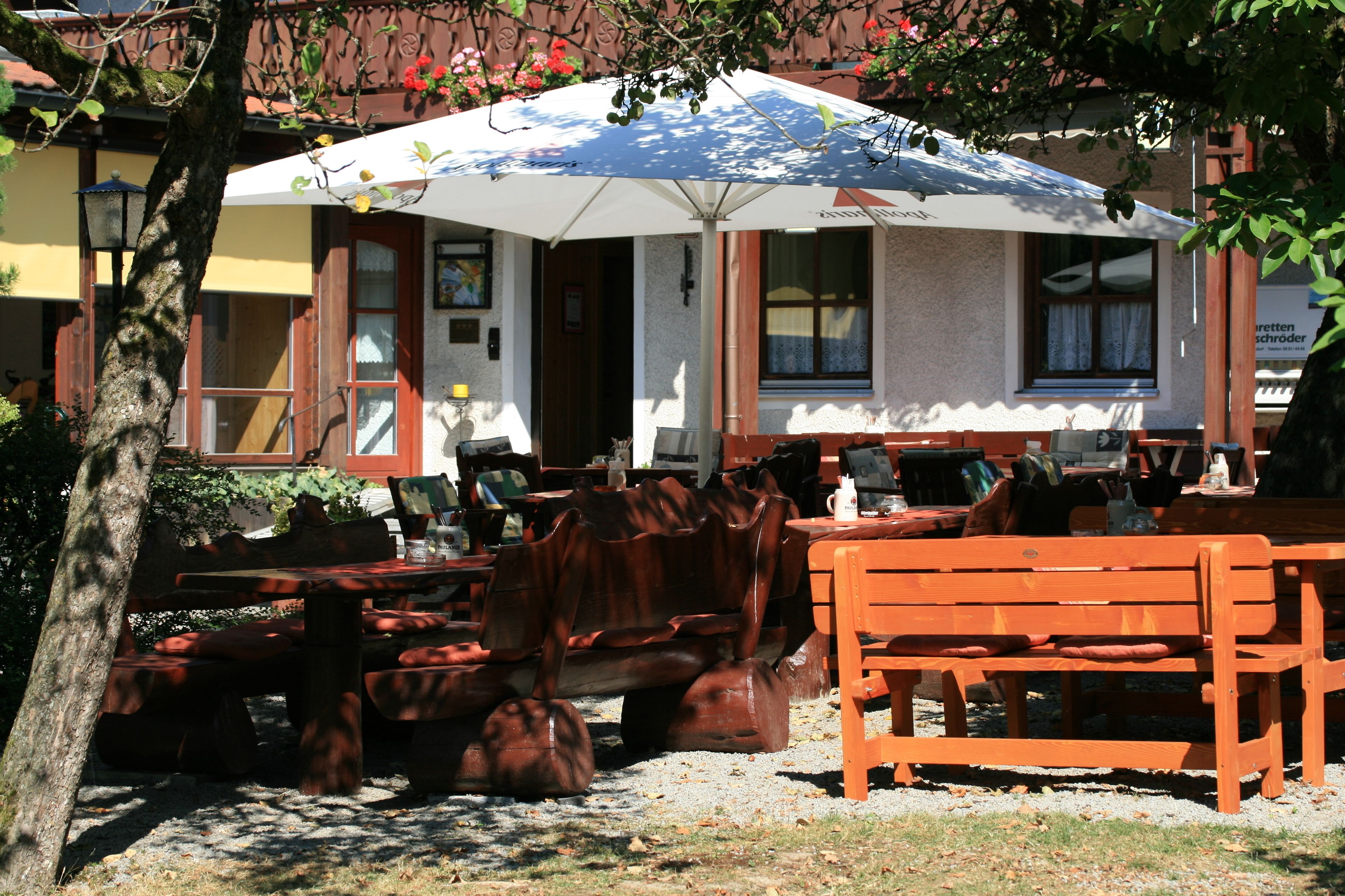 An outdoor seating area with wooden benches and tables, a large umbrella providing shade, and a building with a yellow facade in the background.