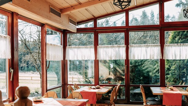 A dining area with wooden tables and chairs, large windows with lace curtains, and a view of trees and a wooden deck outside. - Waldpension Jägerstüberl (Bad Griesbach im Rottal)