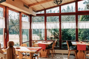 A dining area with wooden tables and chairs, large windows with lace curtains, and a view of trees and a wooden deck outside. - Waldpension Jägerstüberl (Bad Griesbach im Rottal)