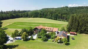 A large house with a red roof, surrounded by green fields and a forested hill. - Waldpension Jägerstüberl (Bad Griesbach im Rottal)
