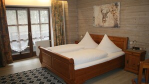 A wooden bed with white linens, a nightstand with a lamp, a window with curtains, and a deer head artwork on the wall. - Waldpension Jägerstüberl (Bad Griesbach im Rottal)