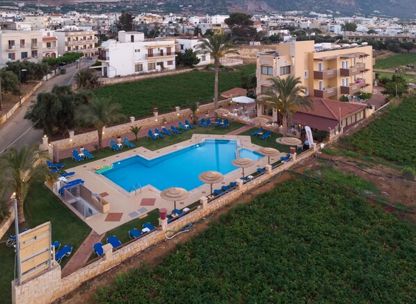 Un resort con piscina rodeada de sillas de descanso y sombrillas, con edificios residenciales y vegetación de fondo.