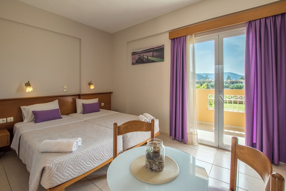 A hotel room with a bed, a table with a glass top, purple curtains, and a view of the outdoors.
