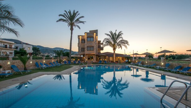 Una piscina con sillones de descanso y palmeras, rodeada de edificios de fachadas de piedra y un paisaje montañoso al fondo.