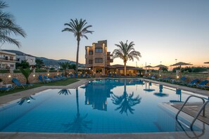 Una piscina con sillones de descanso y palmeras, rodeada de edificios de fachadas de piedra y un paisaje montañoso al fondo.