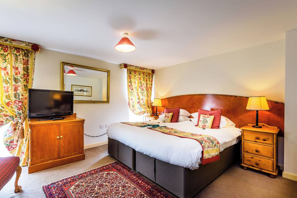 A hotel room with a bed, a television, a wooden cabinet, a nightstand with a lamp, and a patterned rug.