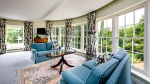 A spacious living room with a blue sofa, a matching armchair, and a coffee table. Large windows with floral curtains offer a view of greenery outside. - Rectory Farm (Cambridge)