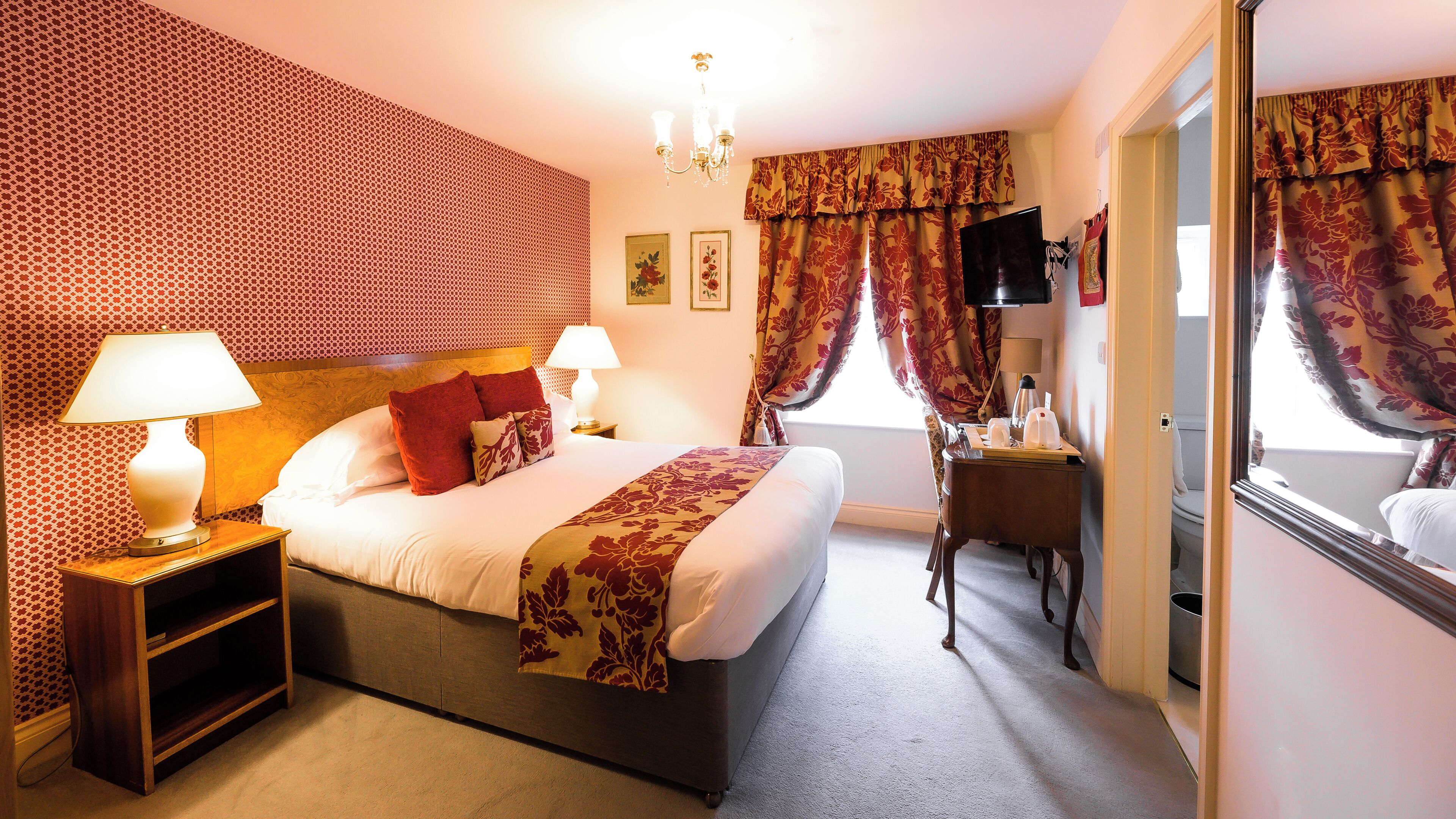 A hotel room with a large bed, two bedside lamps, a desk with a chair, a flat-screen TV, and a window with curtains.