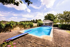 A rectangular swimming pool with a blue edge, surrounded by stone paving, and a small building with a gabled roof in the background.