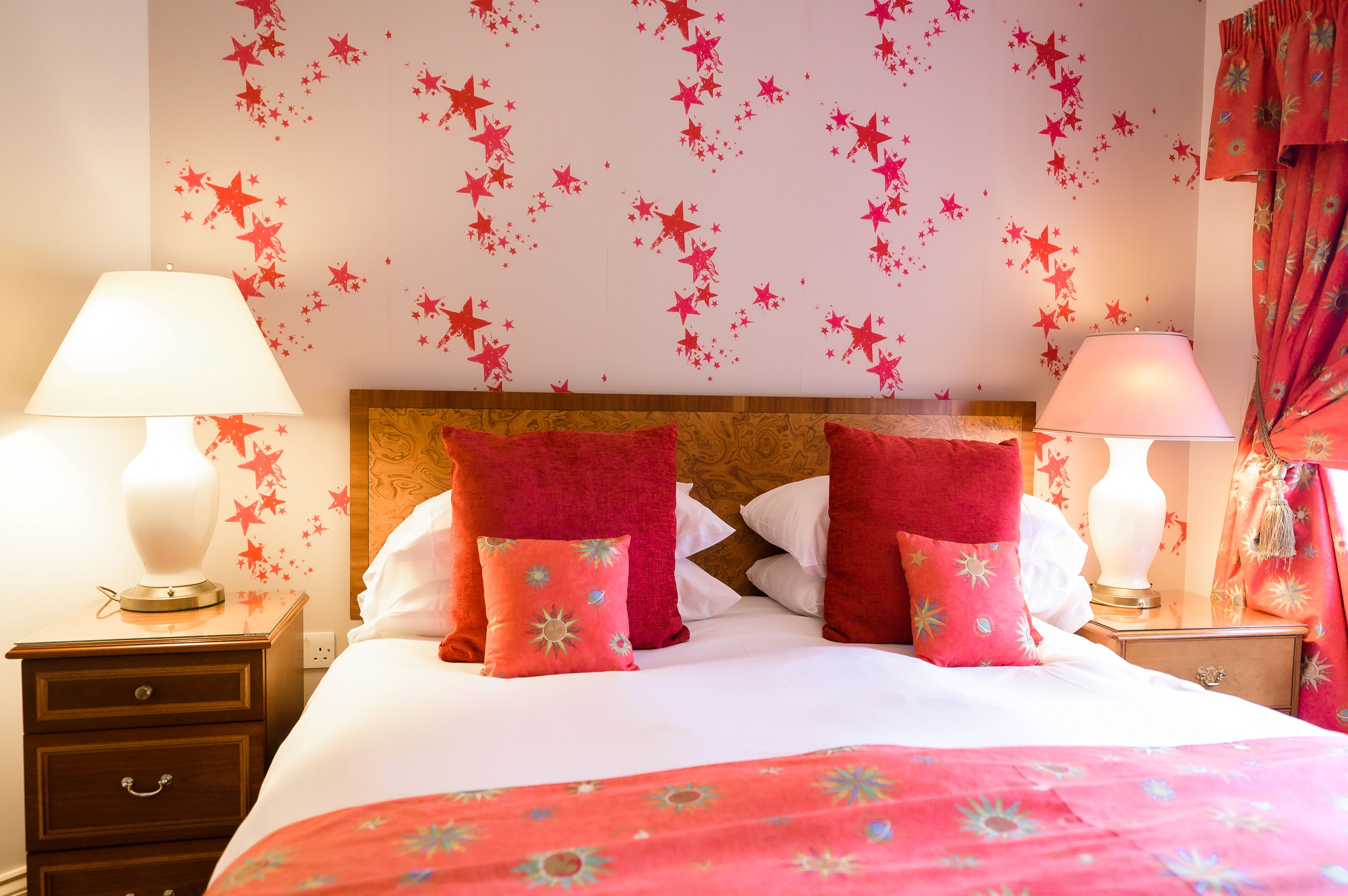 A neatly made bed with red pillows and a pink bedspread, a wooden headboard, and a wall decorated with red star decals.