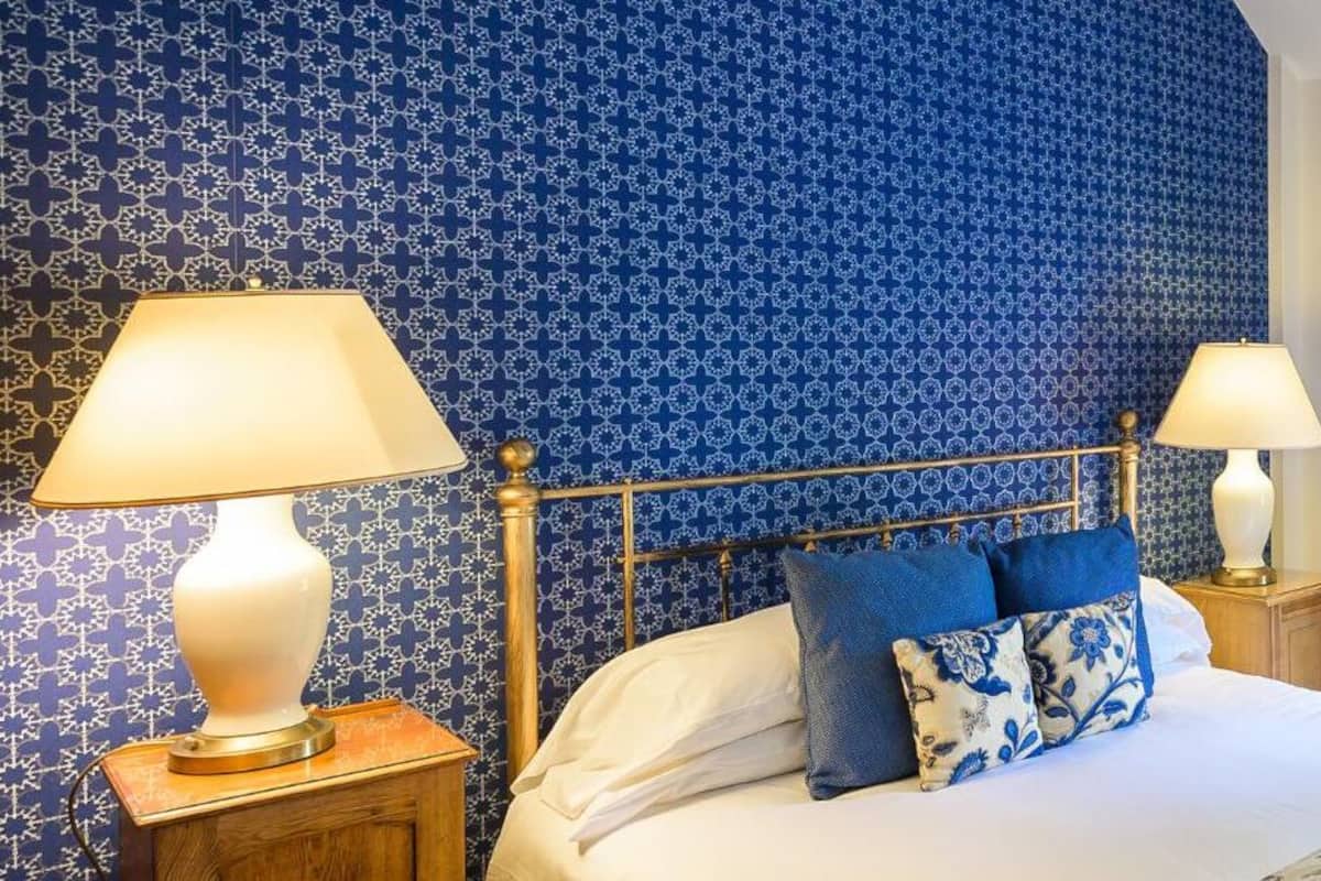 A bedroom with a patterned blue wallpaper, a bed with white and blue bedding, two bedside lamps, and a window with floral curtains.