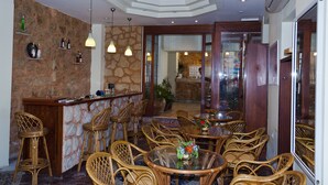 A restaurant with a stone wall, wooden bar, wicker chairs, and a glass table with a vase. - Hotel Krystal (Sitia)