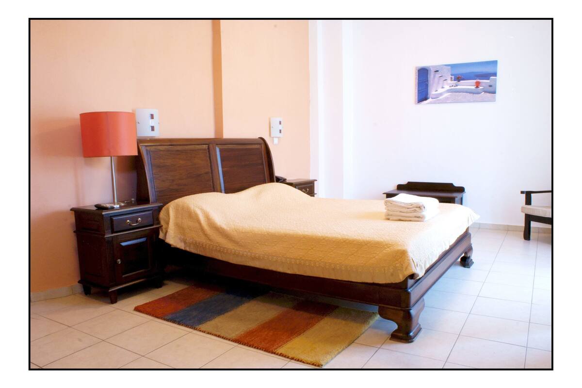 A neatly made bed with a beige blanket, a wooden nightstand with a lamp, and a small table with a chair.
