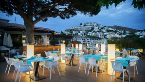 Un espacio al aire libre para comer con sillas y mesas blancas, un árbol grande y vistas a un pueblo en la ladera.