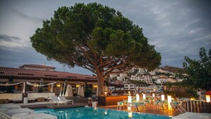 Un área junto a la piscina con sillones de descanso, un árbol grande y vistas a un pueblo en la ladera.