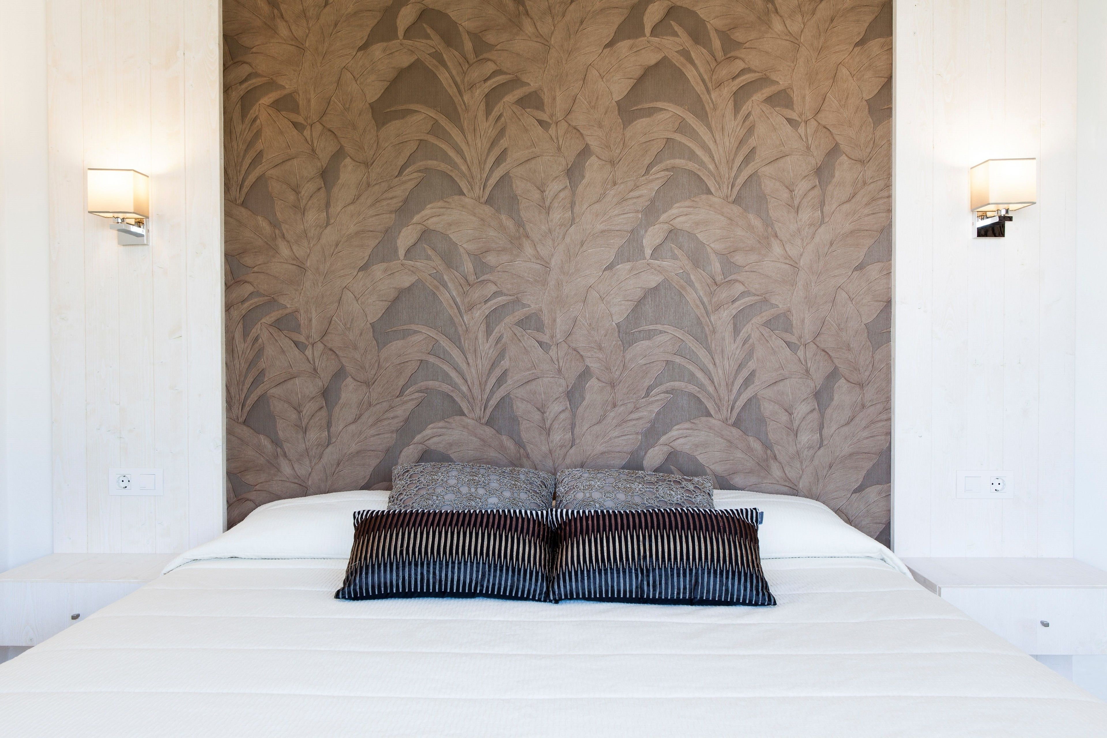 A bed with a patterned wallpaper, two wall-mounted lights, and a pillow.