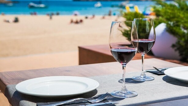 Mesa con dos copas de vino y un plato, con vistas a una playa con gente y barcos.