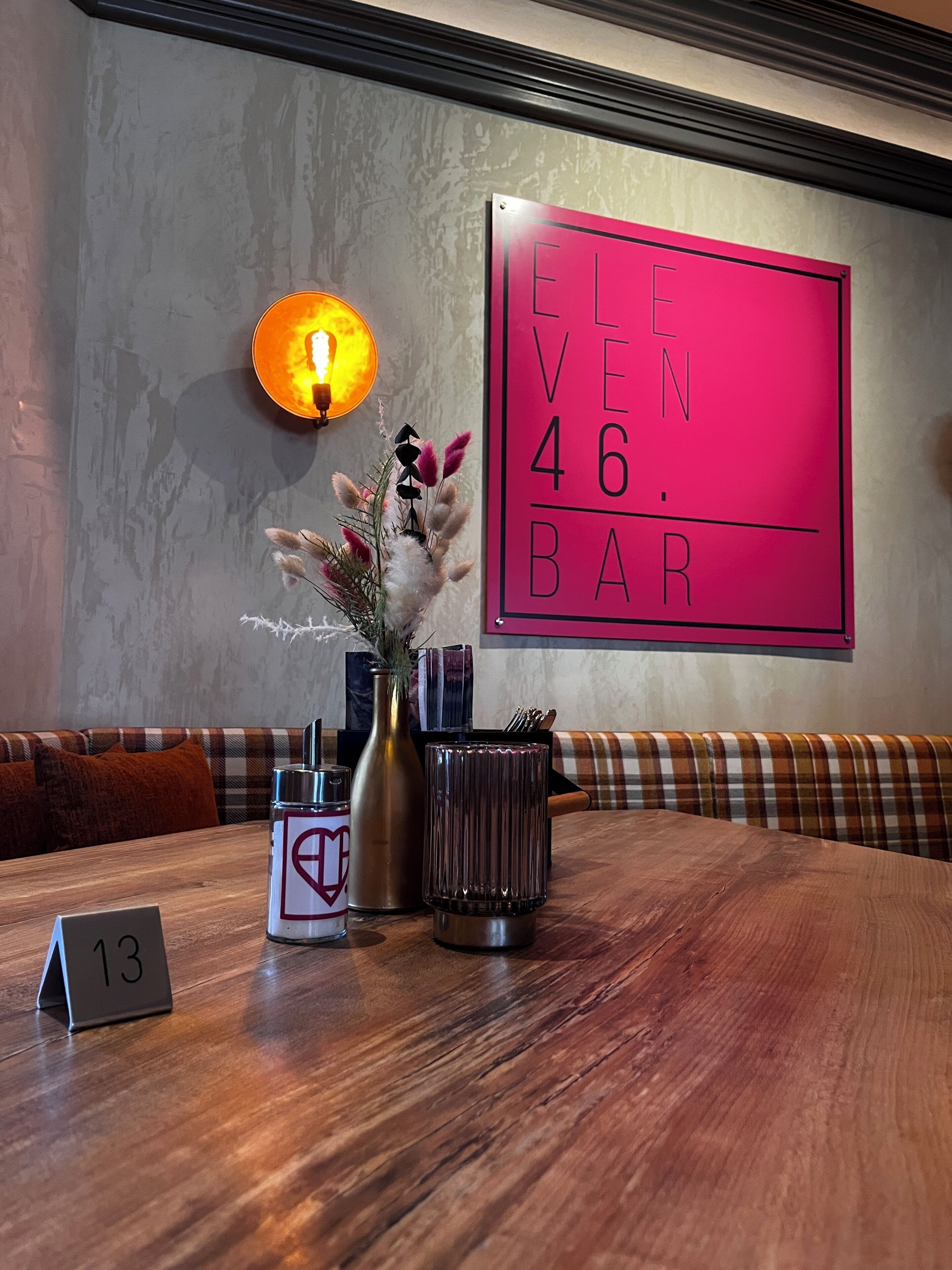A wooden table with a vase of flowers, a candle, and a sign reading 'ELEVEN 46 BAR'.