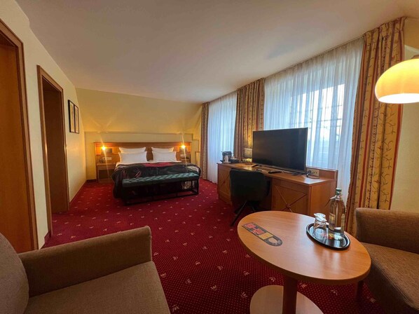 A hotel room with a bed, a desk with a TV, a small table with a bottle, and two chairs. - Das Seela Braunschweig (Braunschweig)