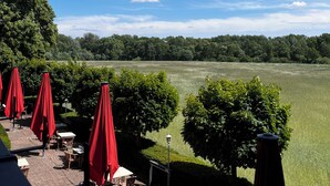 A patio with outdoor seating and red umbrellas, overlooking a green field and trees under a clear blue sky. - Das Seela Braunschweig (Braunschweig)