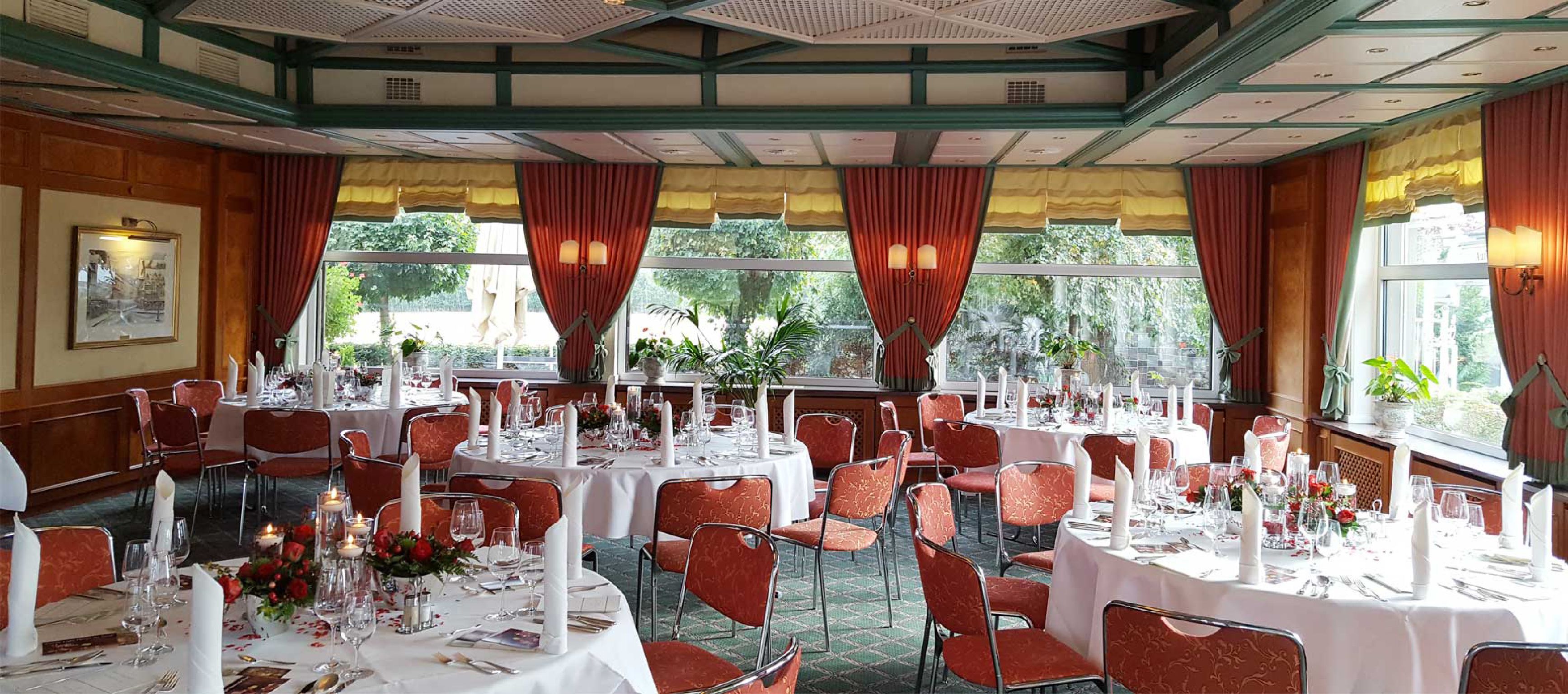 A banquet hall with round tables set for dining, red upholstered chairs, and large windows with curtains.