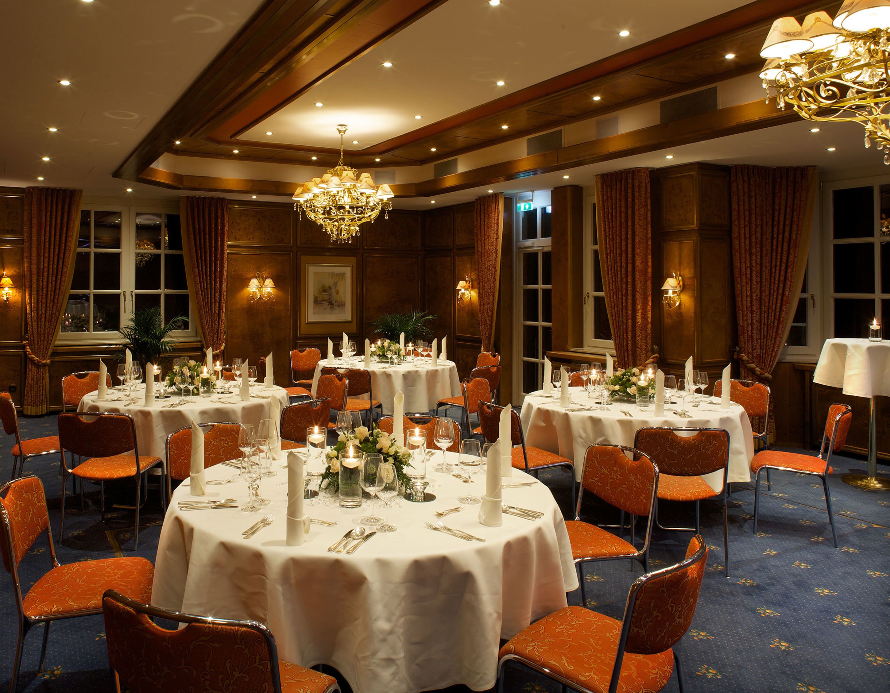 A formal dining room with round tables set for multiple guests, featuring white tablecloths, candles, and orange upholstered chairs.