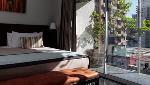 A modern hotel room with a large bed, a bench, a potted plant, and a city view through large windows. - Gen Rooms (Santiago)