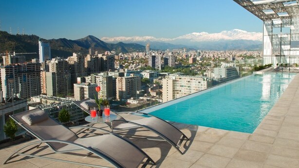A rooftop pool with lounge chairs and a table, overlooking a cityscape with mountains in the background. - Gen Rooms (Santiago)