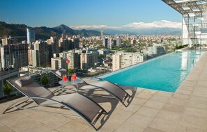 A rooftop pool with lounge chairs and a table, overlooking a cityscape with mountains in the background. - Gen Rooms (Santiago)
