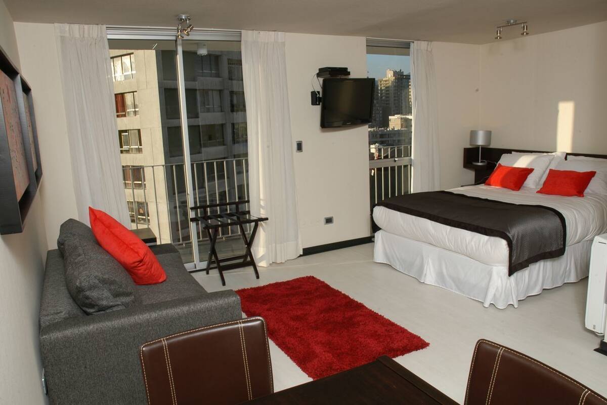 A hotel room with a bed, a sofa, a TV, and a balcony with a view of buildings.