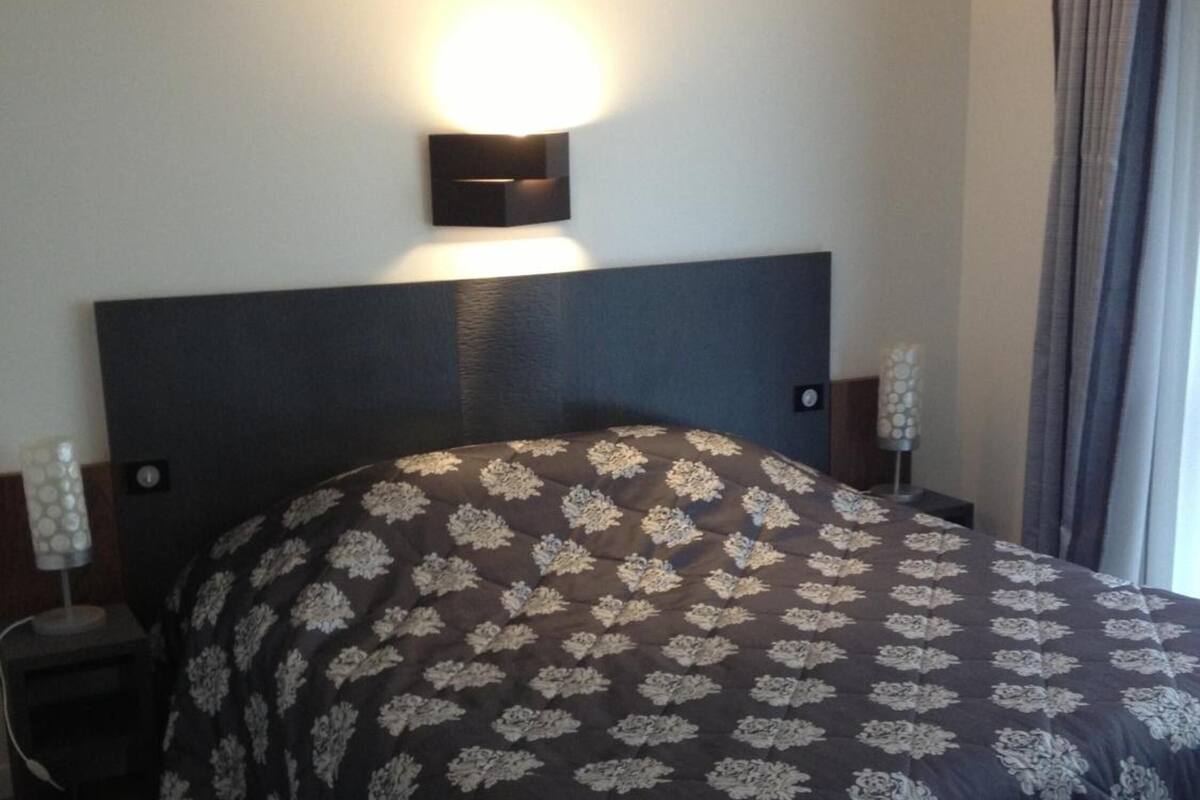 A hotel room with a large bed, a nightstand, a wall-mounted lamp, and a window with curtains.