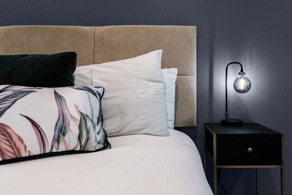 A bed with a headboard, pillows, a nightstand with a lamp, and a wall. - Regent Hotel (Cambridge)
