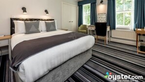 A hotel room with a large bed, a desk, and a chair. There is a window with curtains and a view of greenery outside.