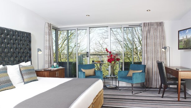A modern hotel room with a large bed, a desk, two chairs, and a view of the outdoors.