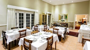 A dining area with multiple tables set for meals, large windows with curtains, and a view of the outside.
