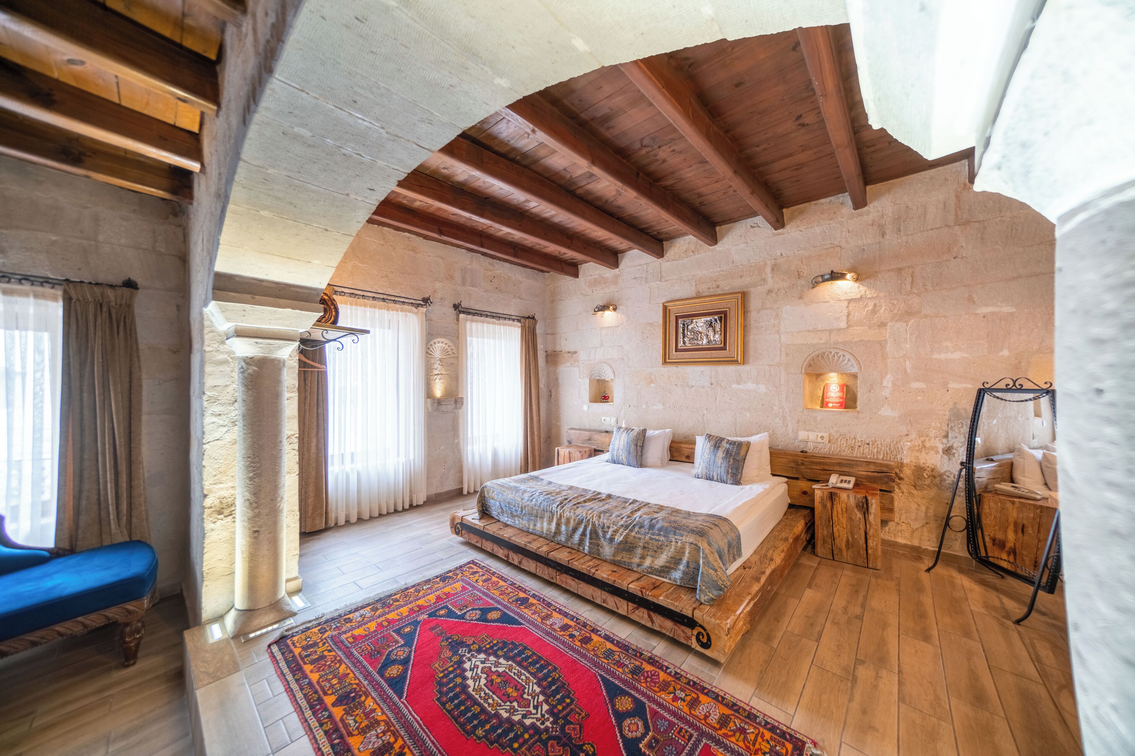 A bedroom with a large bed, a wooden bench, a patterned rug, stone walls, and a stone fireplace.