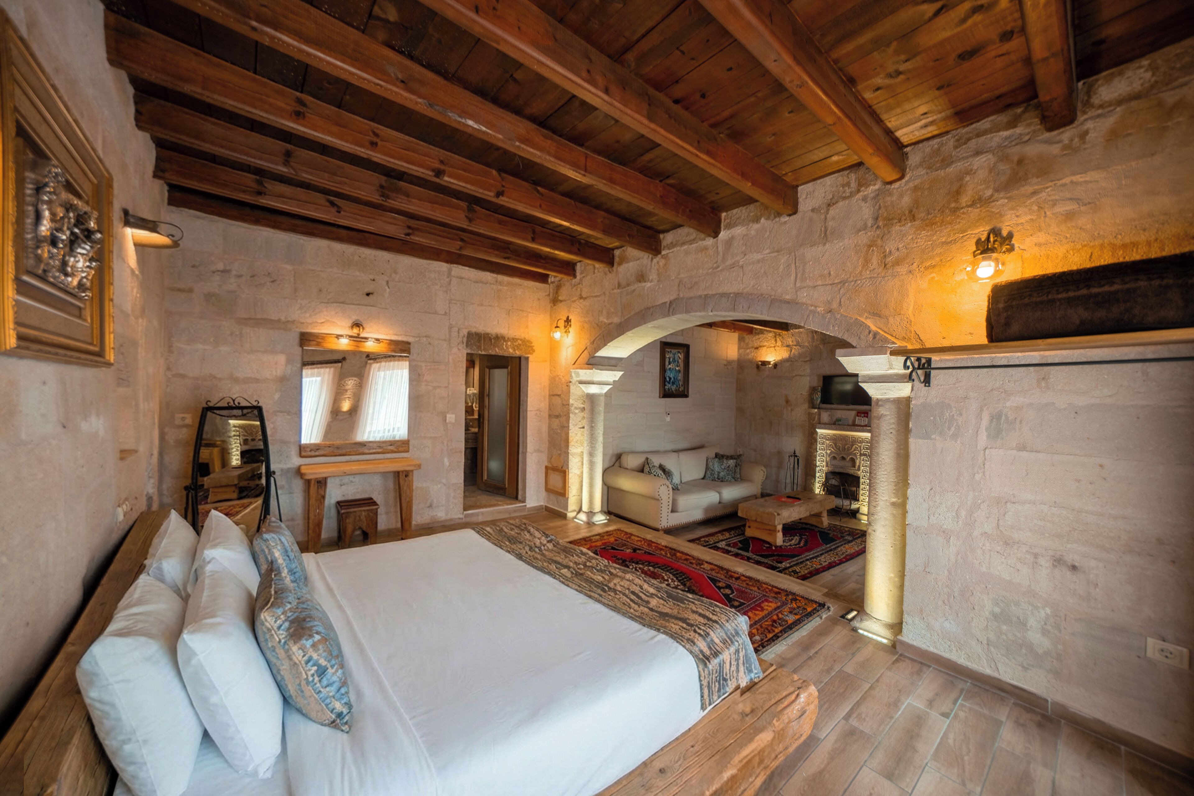 A rustic bedroom with a wooden ceiling, stone walls, a bed with pillows, a sofa, and a fireplace.