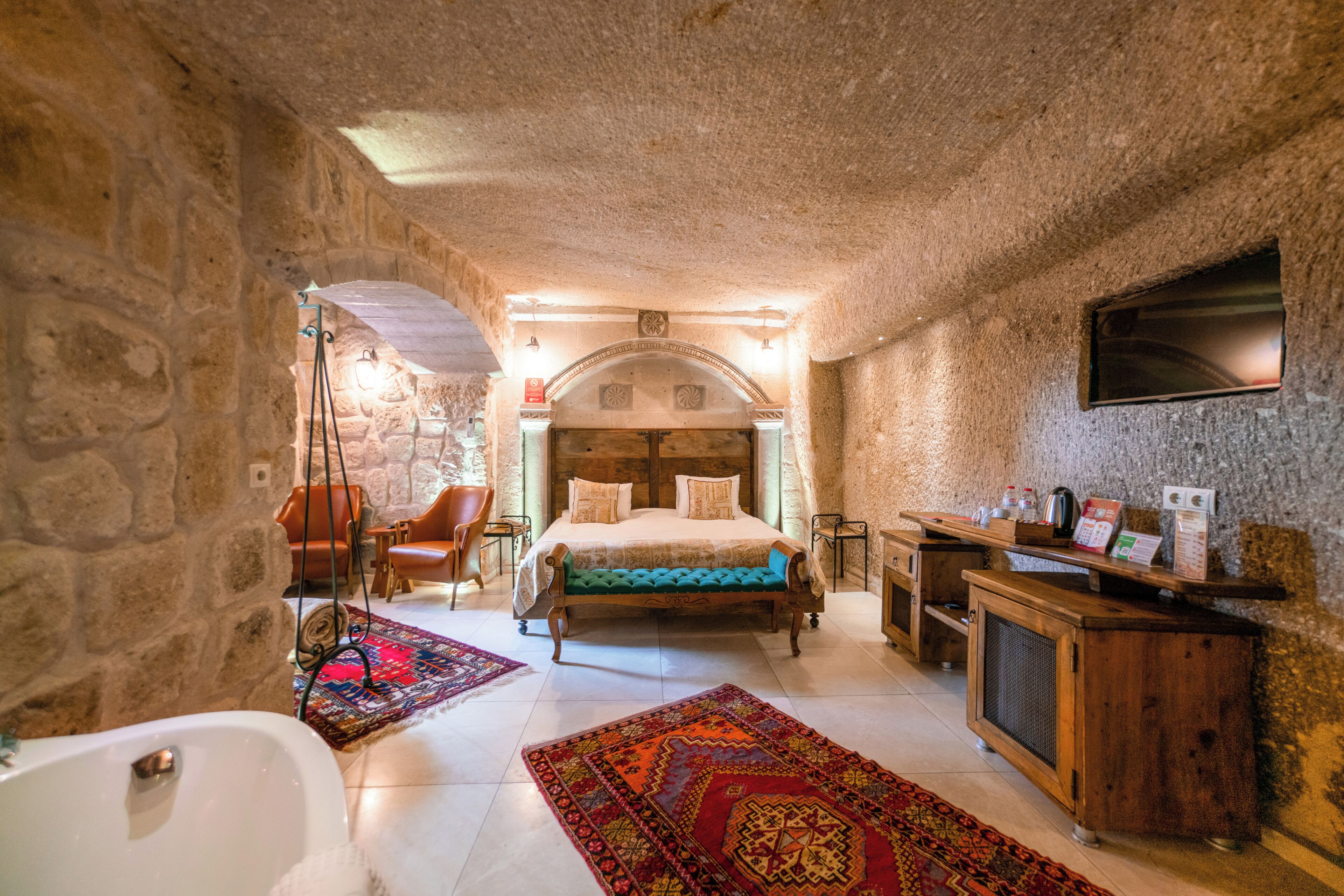 A stone-walled bedroom with a bed, a wooden dresser, a desk, and a television on the wall.