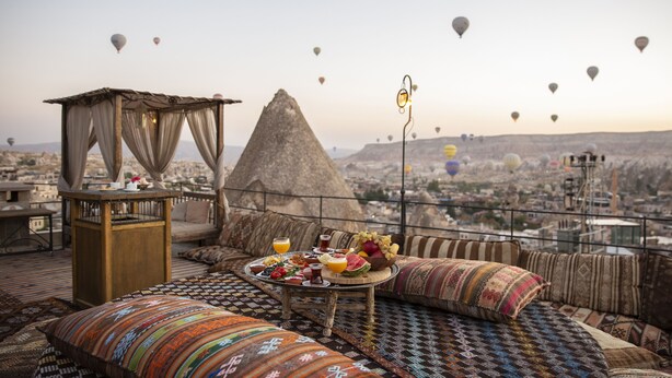 Una terraza en la azotea con una mesa lista para el desayuno, una lámpara decorativa y vistas de la ciudad con globos aerostáticos a lo lejos.