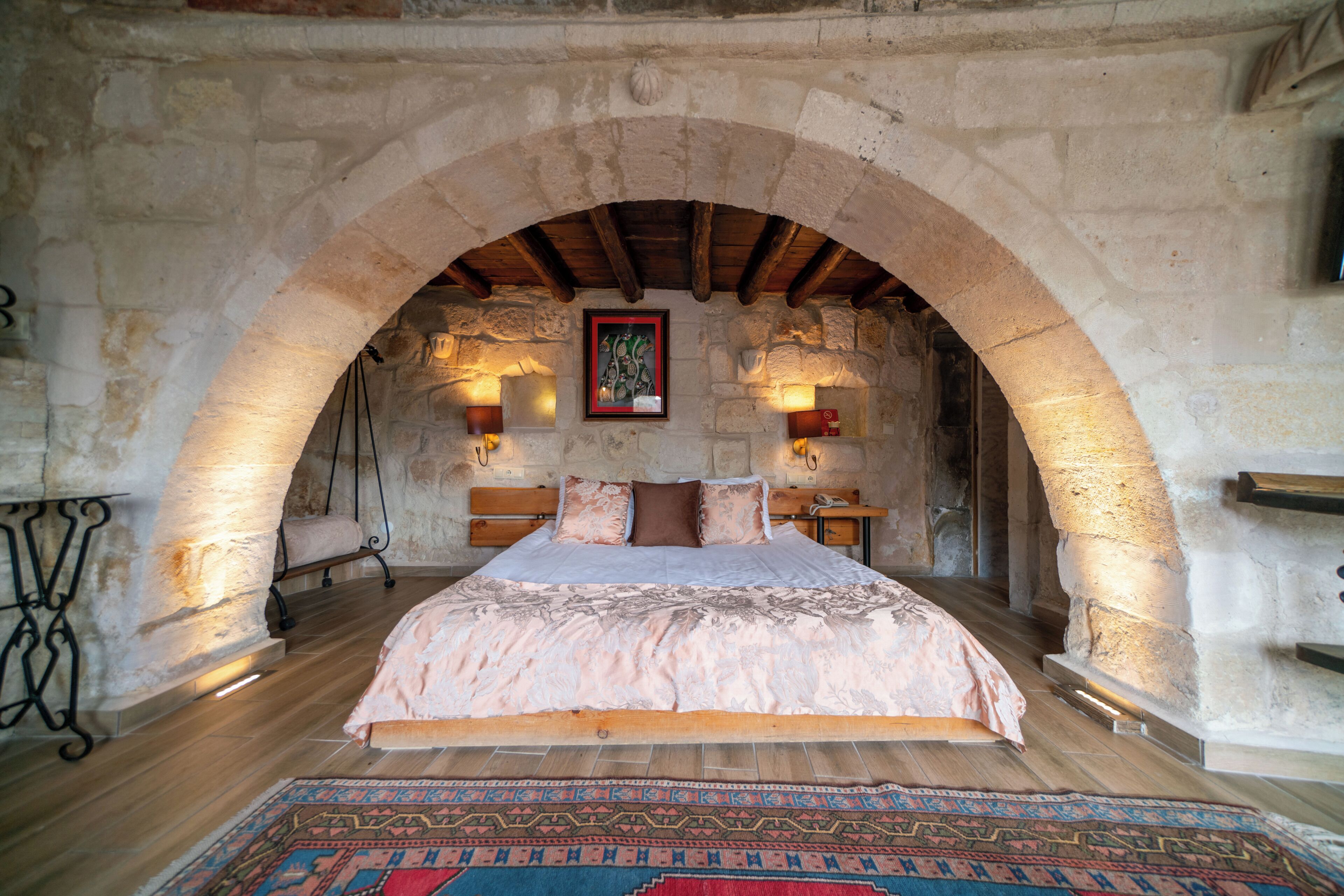Un dormitorio con un arco de piedra, una cama con colcha estampada, una alfombra y dos lámparas fijadas a la pared.