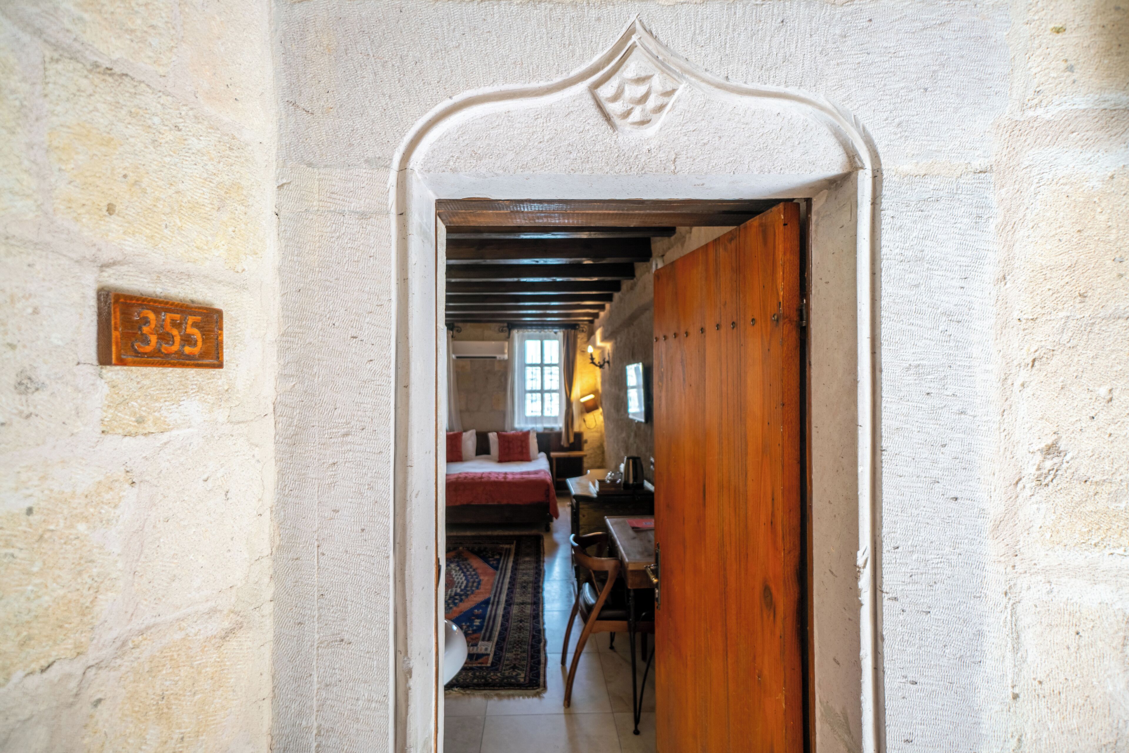 An interior hallway with a wooden door, a number plaque, a bed, a desk, and a window.