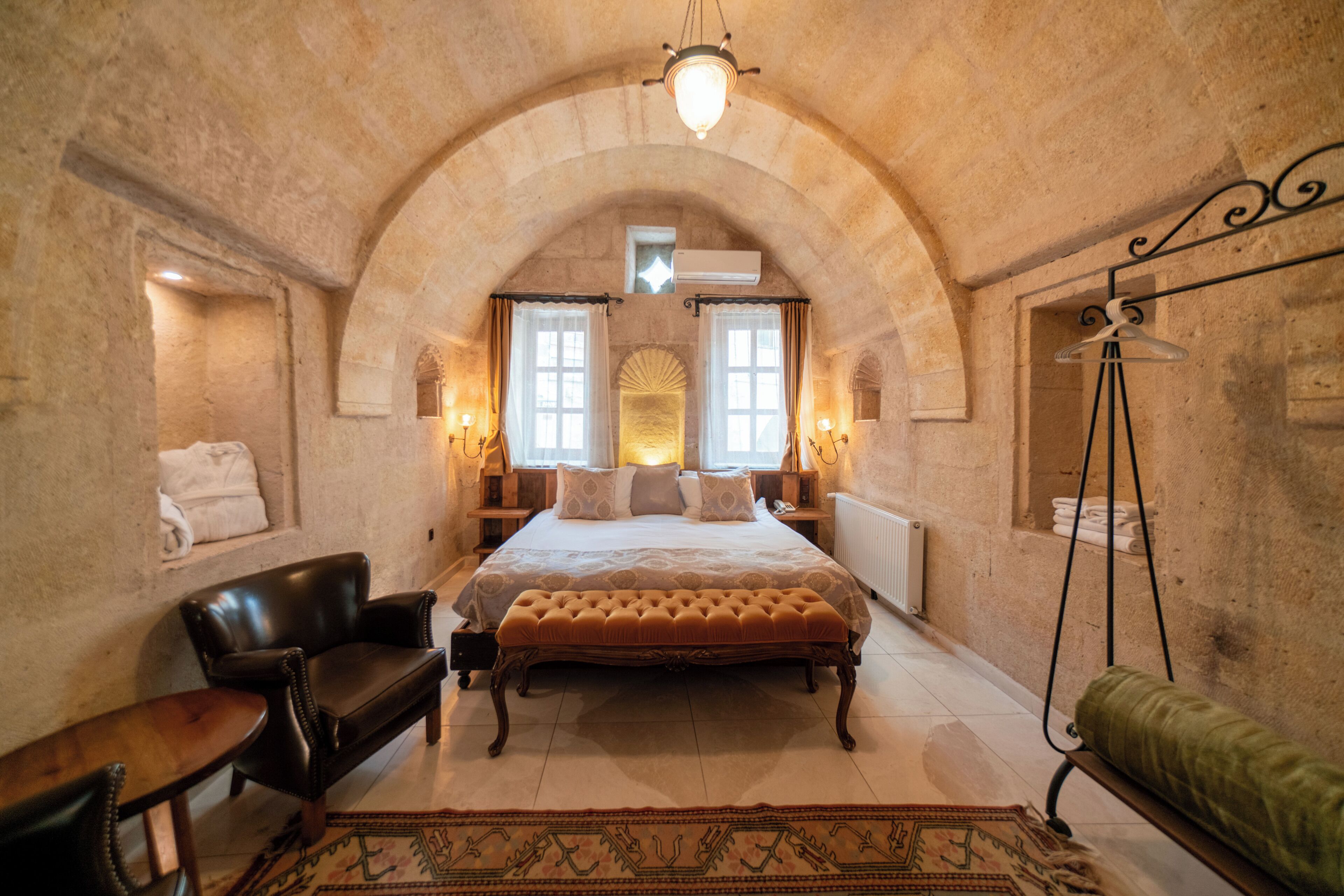 A bedroom with a large bed, a hanging light fixture, a bench, and a radiator.