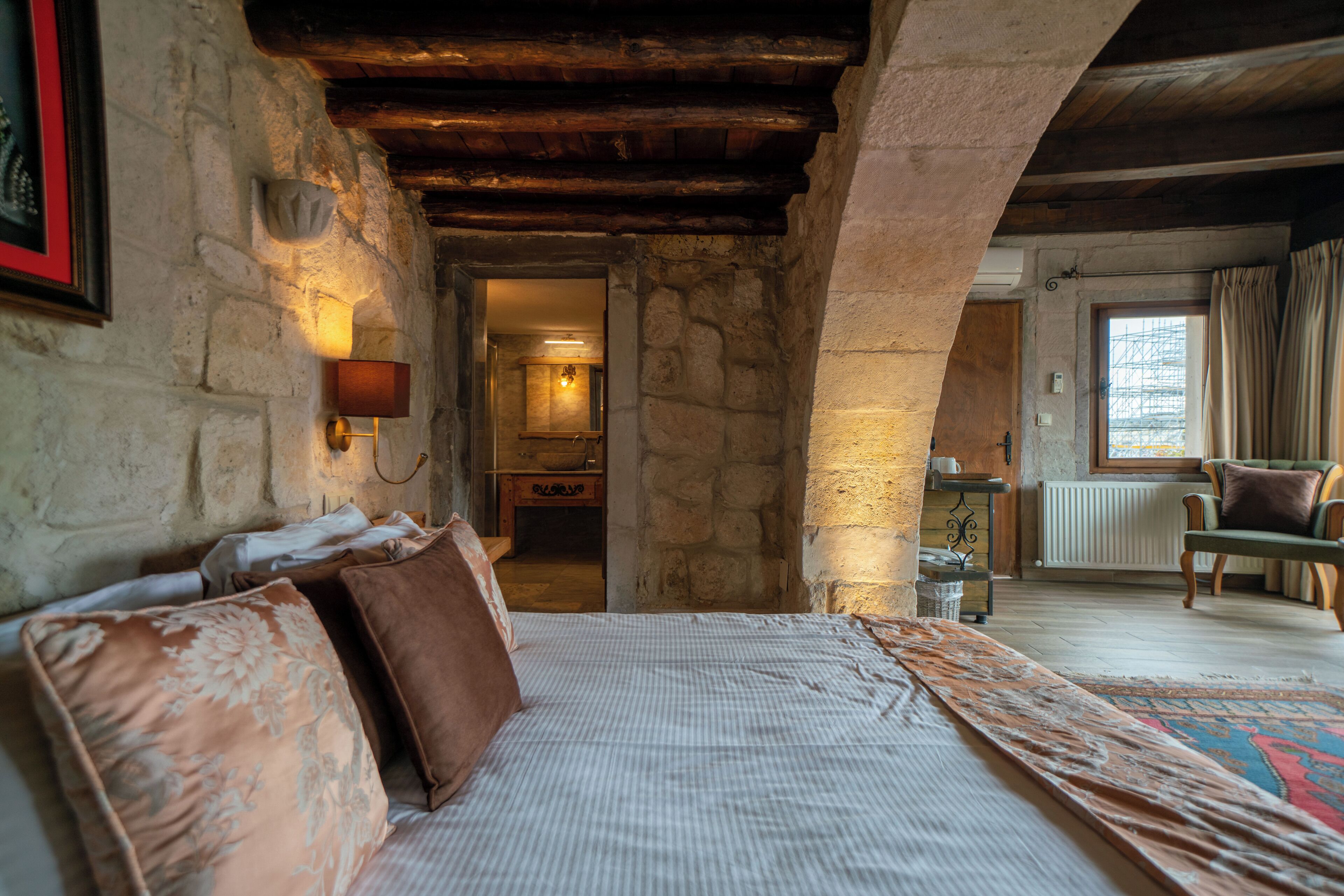 A rustic room with stone walls, a wooden ceiling, a bed with patterned pillows, a small table, and a view through an archway to another room.
