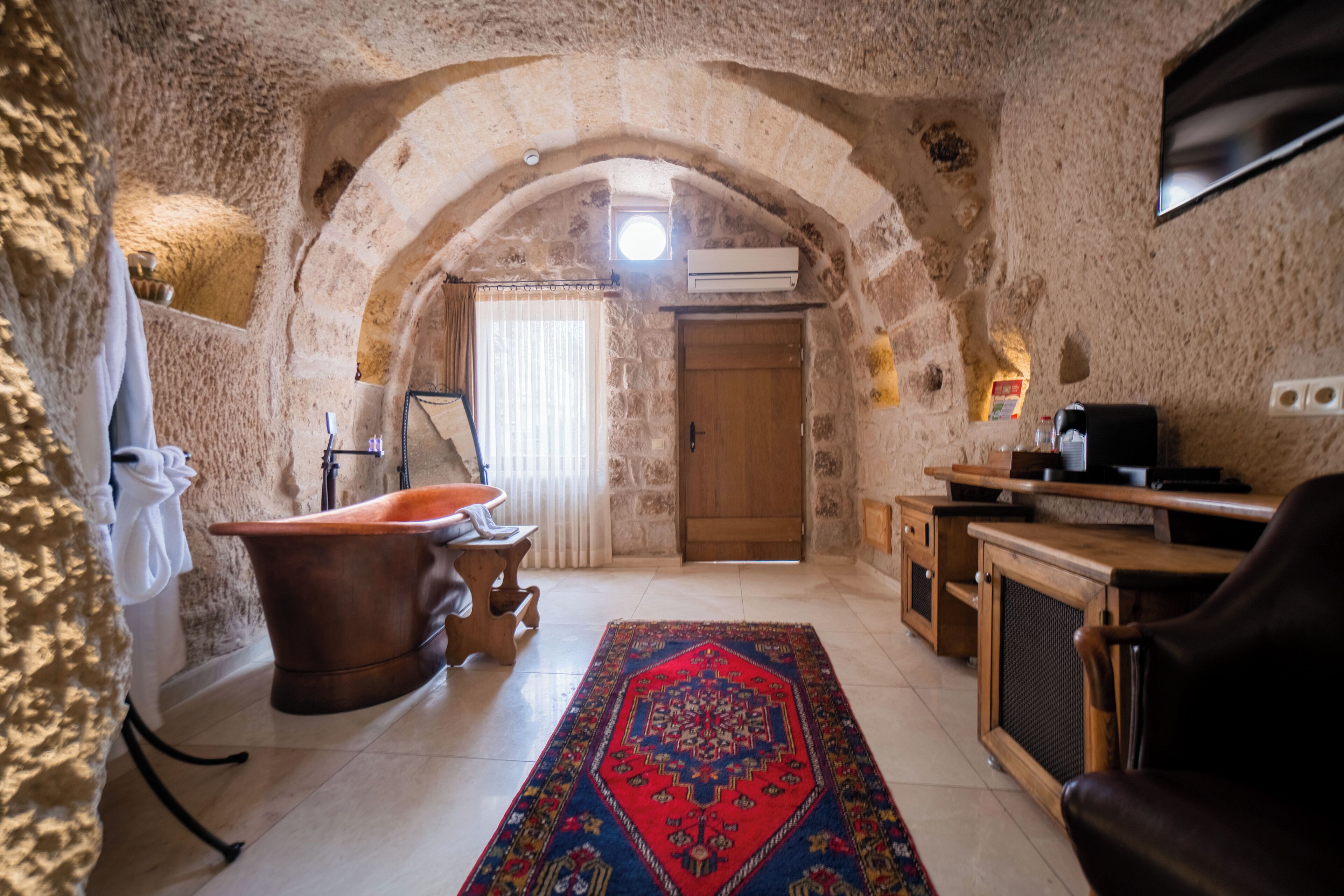 A stone-walled room with a wooden door, a bathtub, a rug, and a TV.