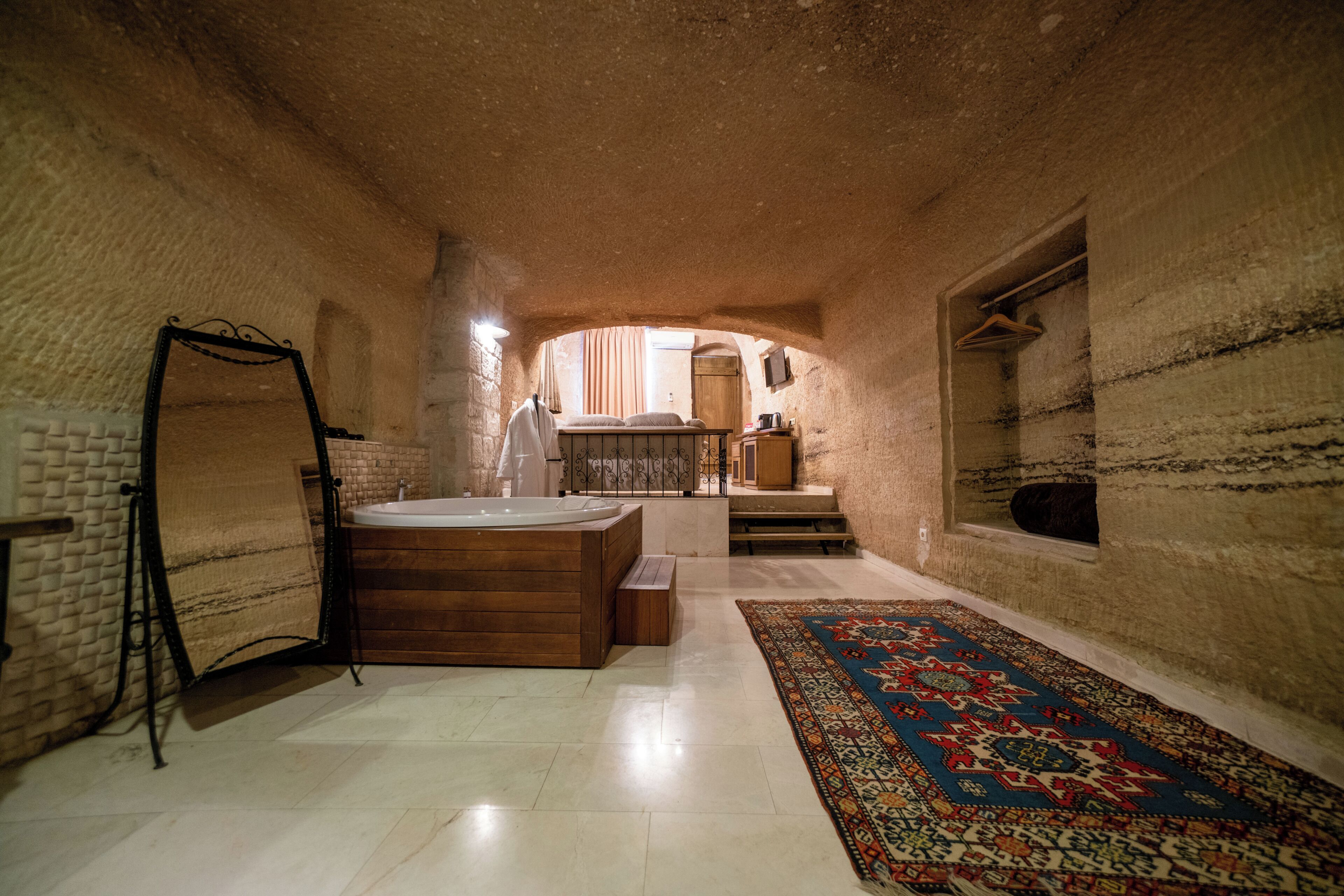 A stone-walled room with a wooden bathtub, a hanging bathrobe, and a patterned rug.